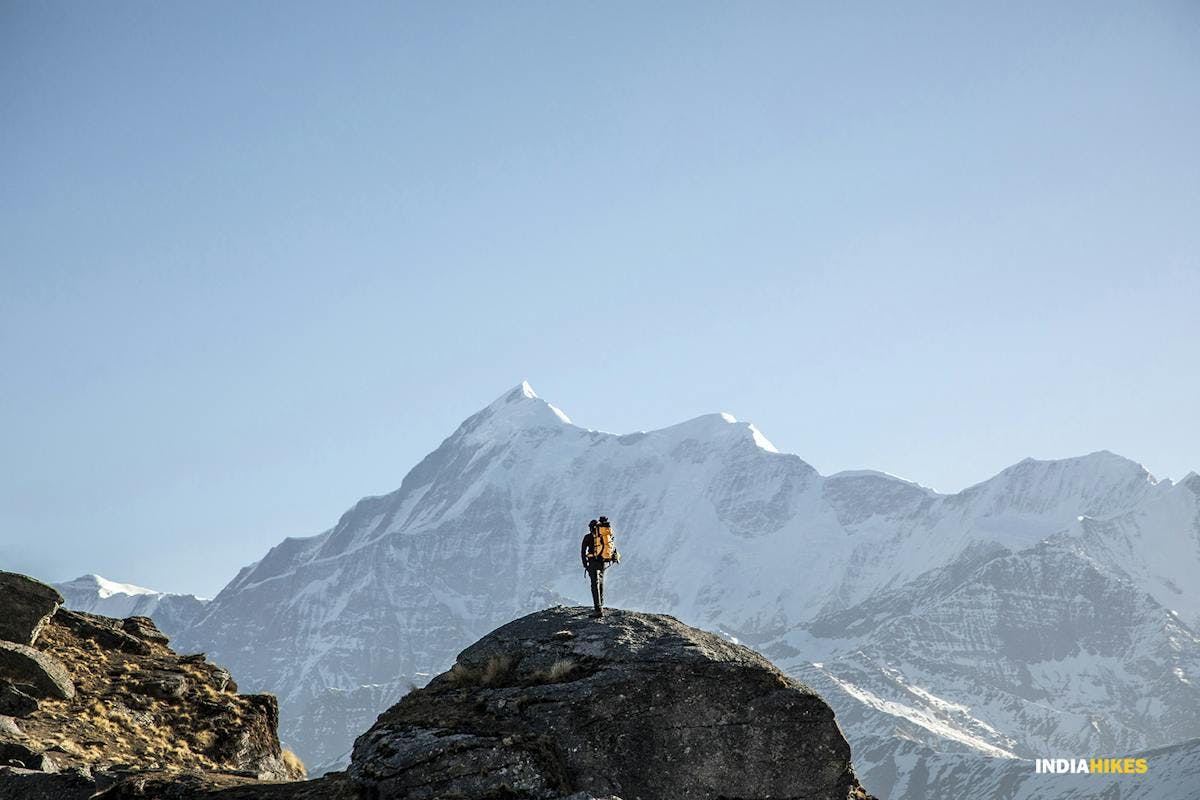 A cinematic photo of a trekker standing on top of a big boulder with Mount Nanda Ghunti in the background
